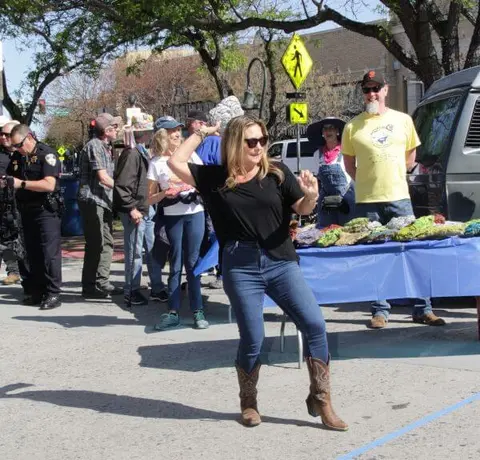 A woman dances outdoors in cowboy boots near a street market or gathering.