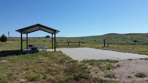 A picnic shelter and concrete pad sit in a grassy area with rolling hills in the background.