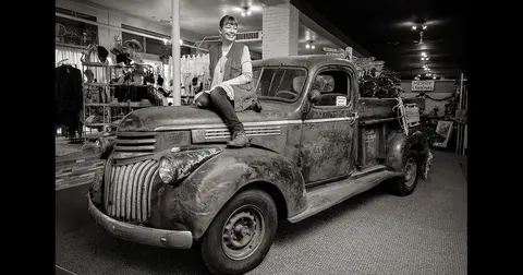 A woman poses sitting on the hood of an old truck indoors.