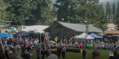 A large crowd gathers outdoors near a rustic building and tents in a wooded area.
