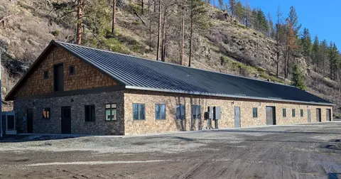 An exterior view of a long stone and shingle building with a metal roof at the base of a rocky hillside.