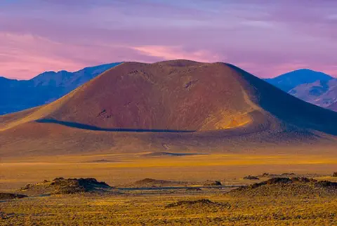 A volcanic crater rises above a desert landscape under a pink twilight sky.