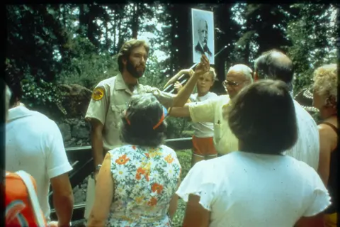 A park ranger holds up a portrait for a group of visitors outdoors.