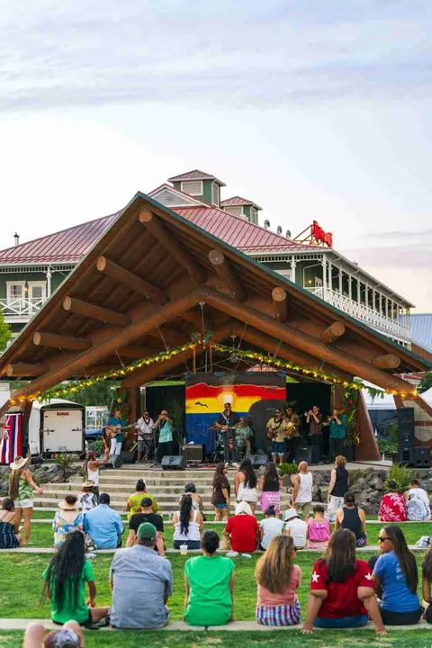 A band performs on an outdoor stage with an audience seated on the lawn.