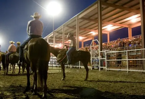 Cowboys on horseback watch an event in a lit arena with a crowd.