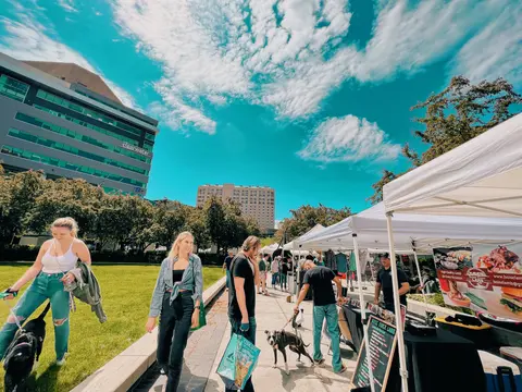 People walk along a pathway lined with vendor tents and a grassy area under a blue sky.