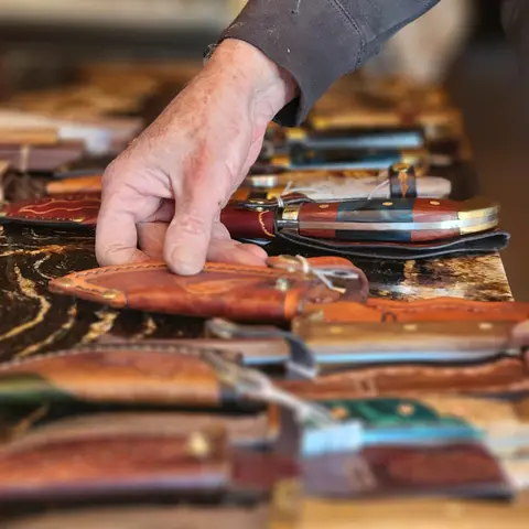 A person's hand reaches for a knife in a leather sheath displayed on a table.