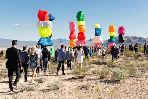 A crowd of people walking in the desert toward tall stacks of brightly painted boulders.