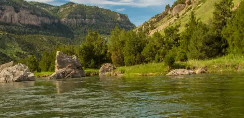 A river flows past large rocks beneath green hills and mountains.
