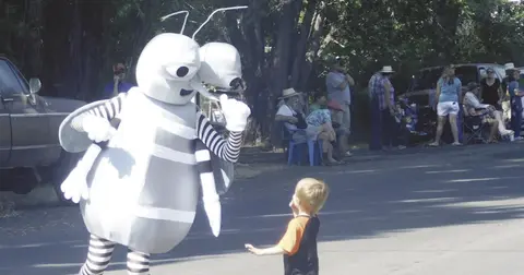 A costumed mosquito mascot interacts with a young child on a street during an outdoor event.