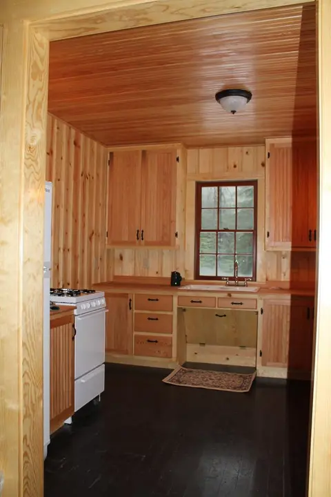 An interior shot of a rustic kitchen paneled in wood with a stove and window.