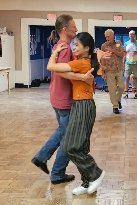 A couple dances inside a hall with wooden floors.