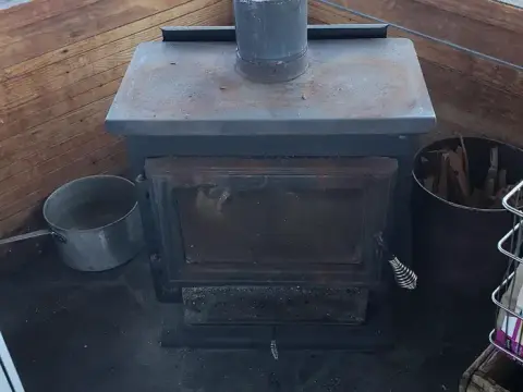 A wood burning stove sits indoors with a pot and firewood nearby.