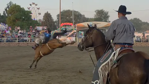 A cowboy rides a bucking horse at a rodeo while another rider watches from the foreground.
