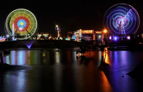 A night view of a carnival with illuminated ferris wheels and lights reflected in water.
