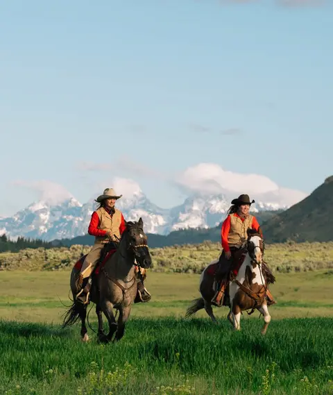 Two women ride horses through a grassy field with mountains in the background.