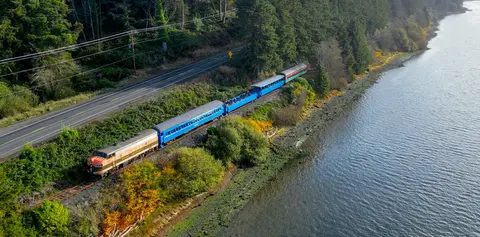 An aerial view of a train traveling along a body of water next to a road and forest.