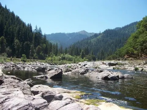 A river flows through a rocky landscape surrounded by forested mountains and blue skies.