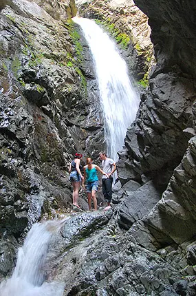 Three people stand near a waterfall in a rocky canyon.