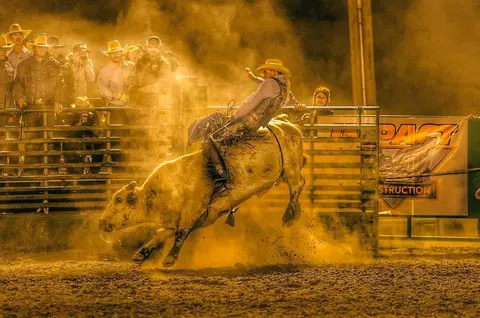 A cowboy rides a bucking bull at a rodeo while spectators watch from behind a fence.