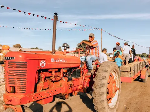 A man drives a red tractor pulling a trailer with people on it.