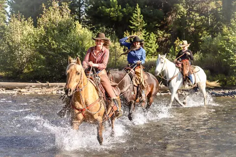 Three people ride horses through a shallow river.