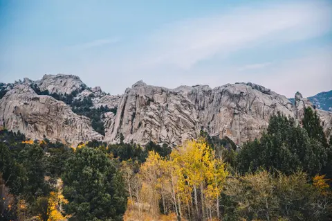 A landscape view of rocky mountains behind green and yellow trees under a blue sky.