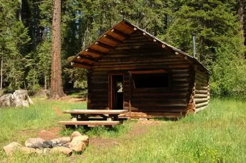 A rustic log cabin stands in a clearing surrounded by trees.