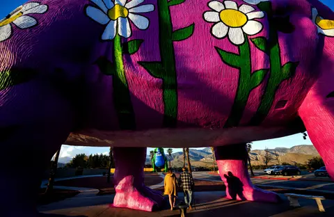 Two people walk underneath a large pink dinosaur sculpture painted with flowers.