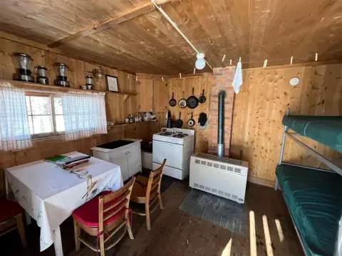 An interior view of a rustic wooden cabin featuring bunk beds, a dining table, and a stove.