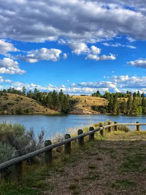 A scenic landscape view of a calm body of water with rolling hills, trees, and a wooden fence under a cloudy blue sky.