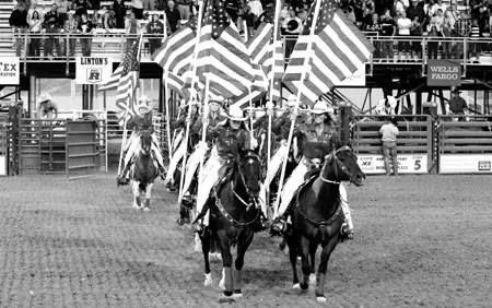 A black and white photo shows horse riders carrying American flags in an arena.