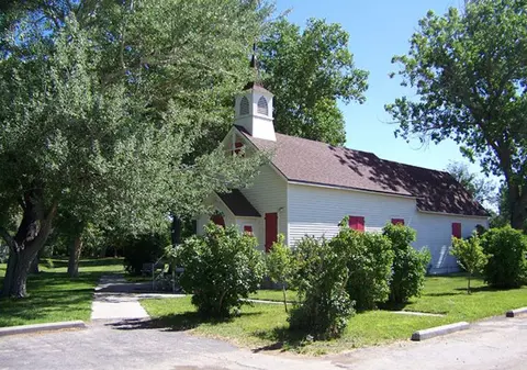 An exterior view of a small white church with a steeple surrounded by trees and bushes.