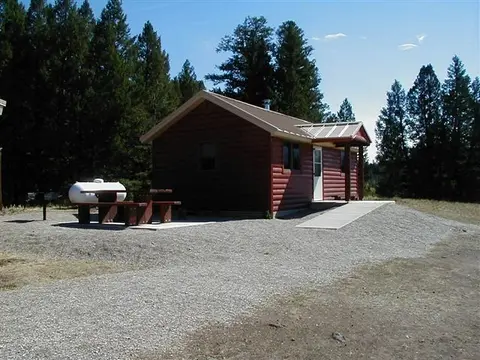 A cabin stands in a wooded area with a picnic table and propane tank outside.