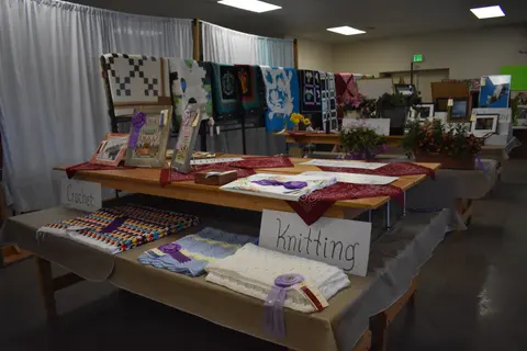 An indoor craft exhibition features tables displaying blankets, quilts, and handmade items with ribbons.