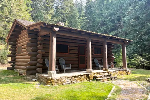 A rustic log cabin stands in a wooded setting with chairs on its front porch.