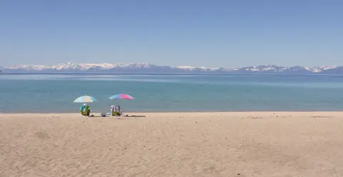 Two beach chairs and umbrellas on a sandy shore facing a large body of water with distant snow-covered mountains.