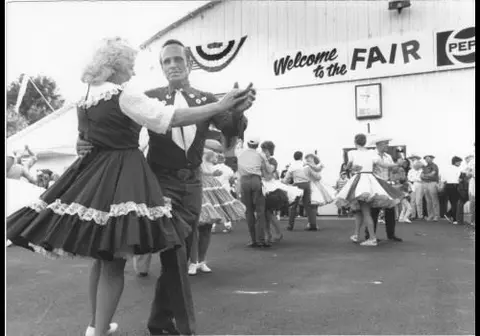 A black and white photograph of people square dancing outside at a fair.