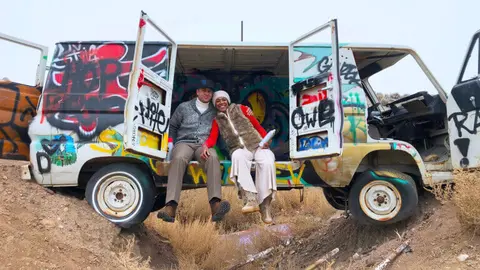 Two people sitting in the open back of a heavily graffiti-covered van.