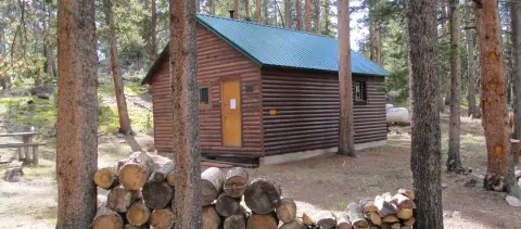 A log cabin sits in a forest with a stack of firewood in the foreground.