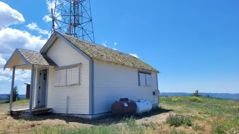 A small white building with a metal tower stands in a grassy landscape under a blue sky.