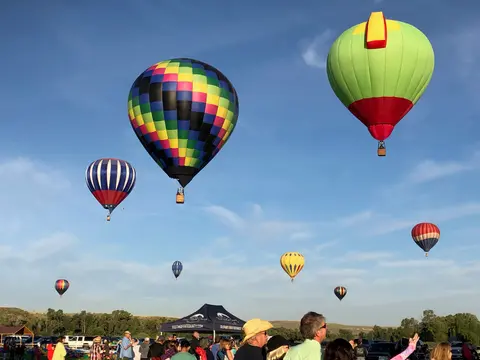 Several hot air balloons fly in a blue sky above a crowd gathered on the ground.