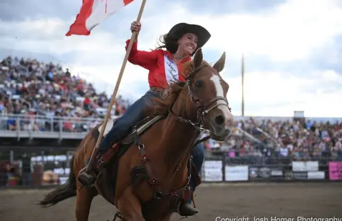 A woman rides a horse carrying a flag in an arena with a crowd.