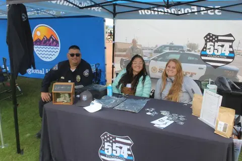 Three people sit behind a booth table set up outdoors under tents.
