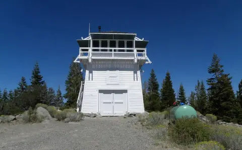 A white wooden fire lookout tower stands under a clear blue sky surrounded by trees.