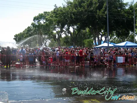A crowd of people gathers behind a barrier as water sprays over a wet street.