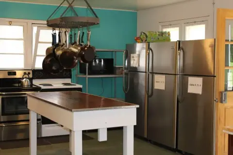 A bright indoor kitchen features a white island table, hanging pots, a stove, and several stainless steel refrigerators.