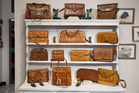 A white shelving unit displays various tooled leather bags and small decorative boots.