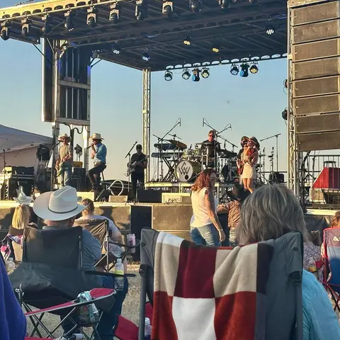 A band performs on an outdoor stage for an audience sitting in lawn chairs.
