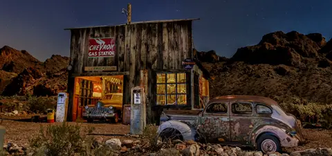 An old wooden gas station at night with vintage cars parked outside and inside.